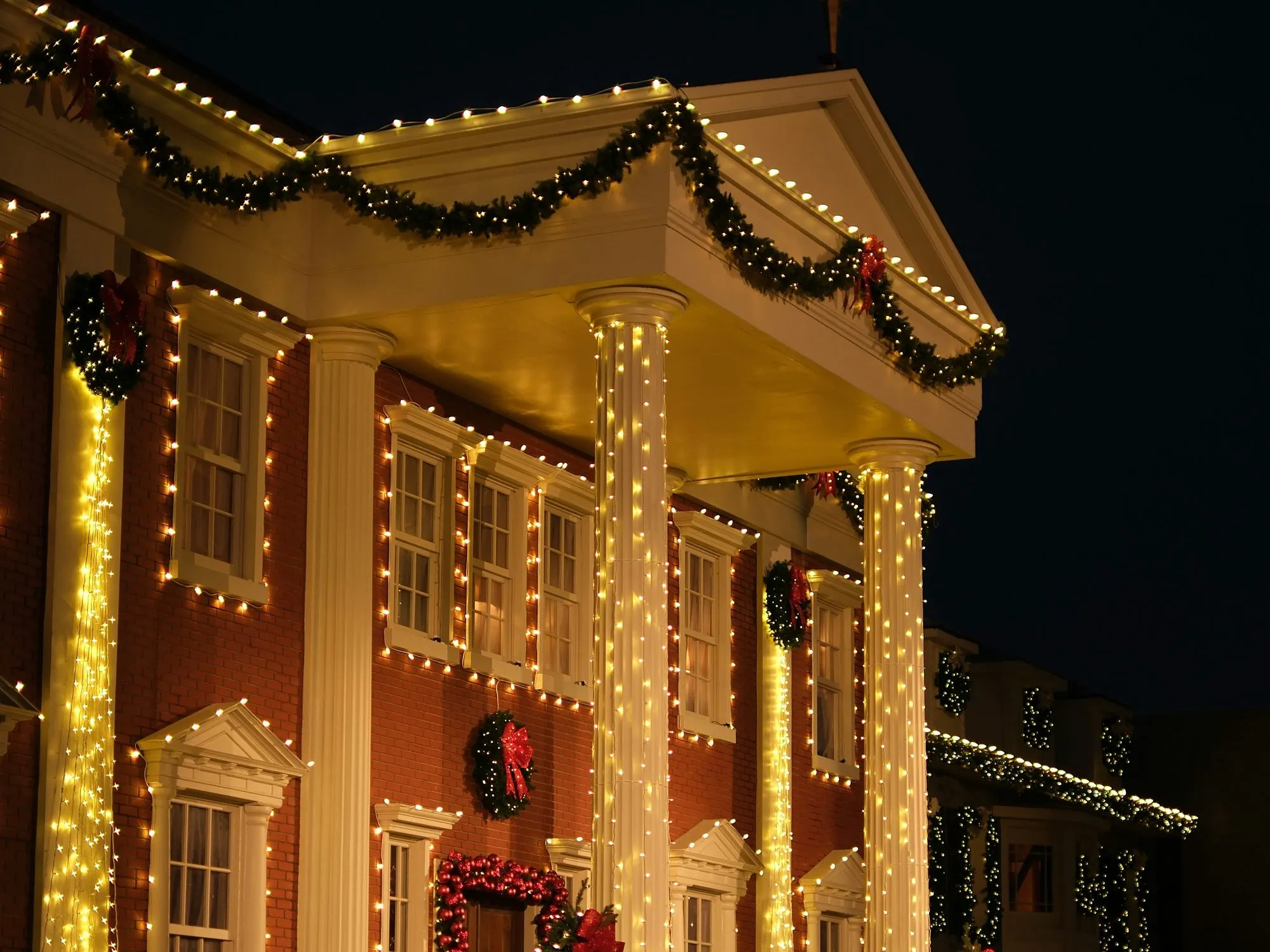 Traditional red brick home with white columns at night, warm white lights along the pediment, windows, and columns, garland and wreaths over the entry